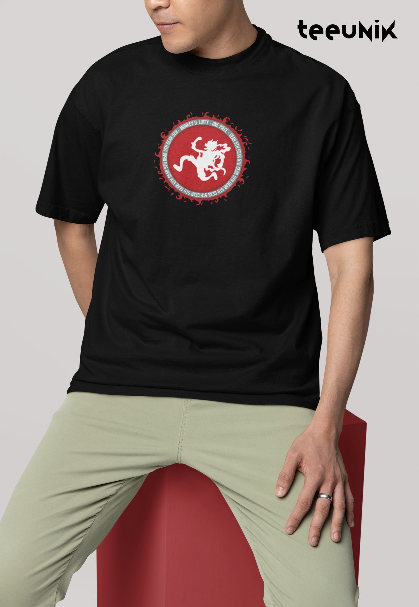 Man sitting on a red box wearing a black tshirt from Teeunik brand that features a front print graphic of Luffy Gear 5 "Nika" with red circle background from One Piece Anime.